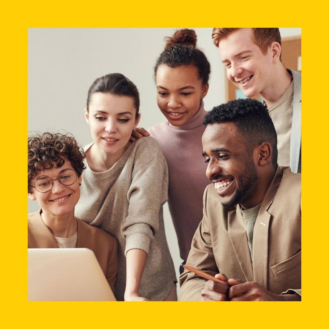 Eine Gruppe von 5 Erwachsenen schauen gemeinsam in ein Laptop und sehen zufrieden aus
